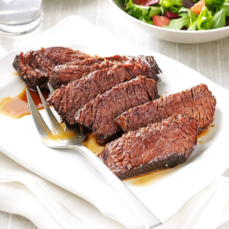 some meat is on a white plate with a fork and bowl of salad in the background