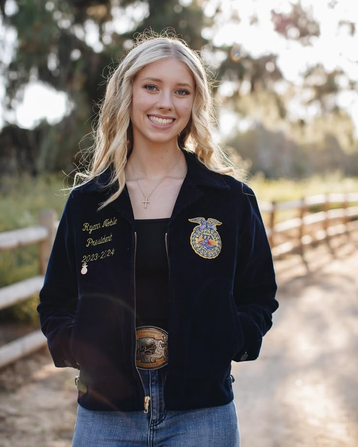 a woman standing in front of a fence wearing a black jacket with patches on it