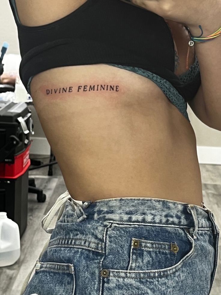 a woman with a tattoo that says divine feminine on her stomach is looking at her cell phone