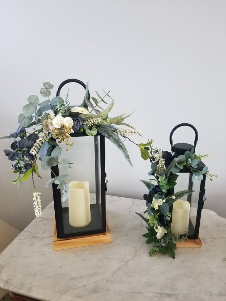 two black lanterns with flowers and candles on a marble table next to a white wall