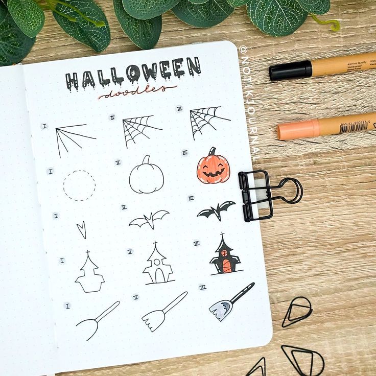 an open notebook with halloween doodles on it next to markers, pencils and leaves