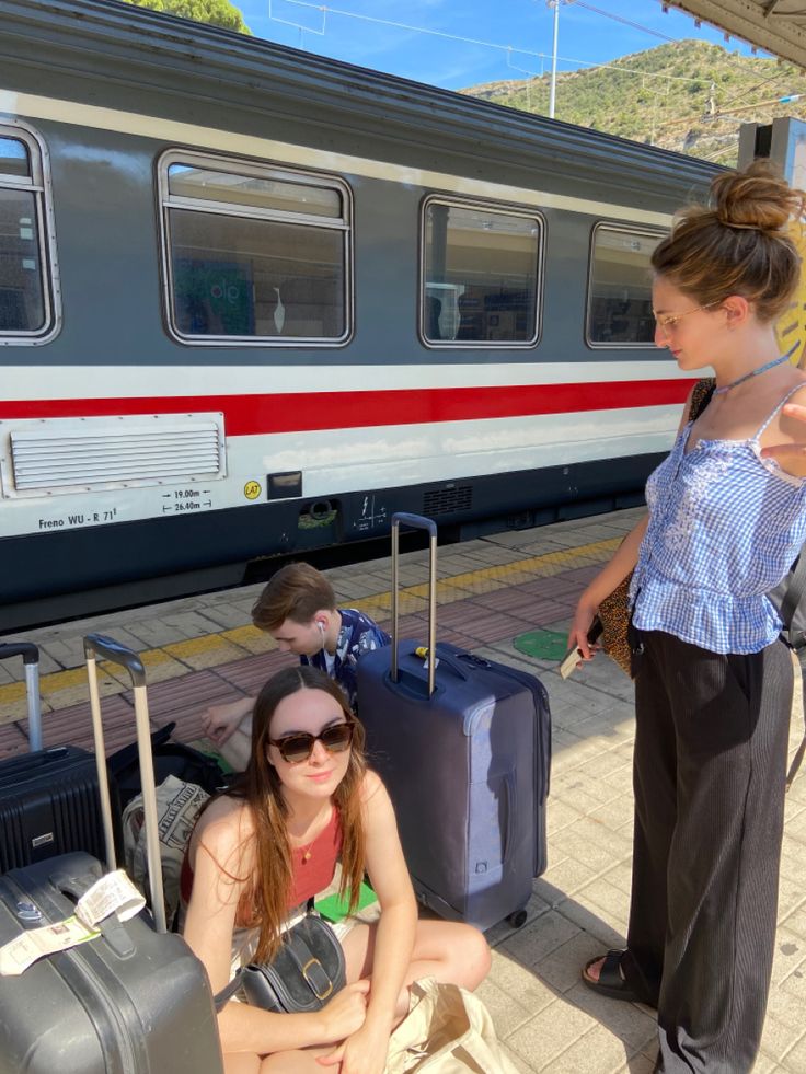 two women sitting on the ground with luggage in front of a train at a station