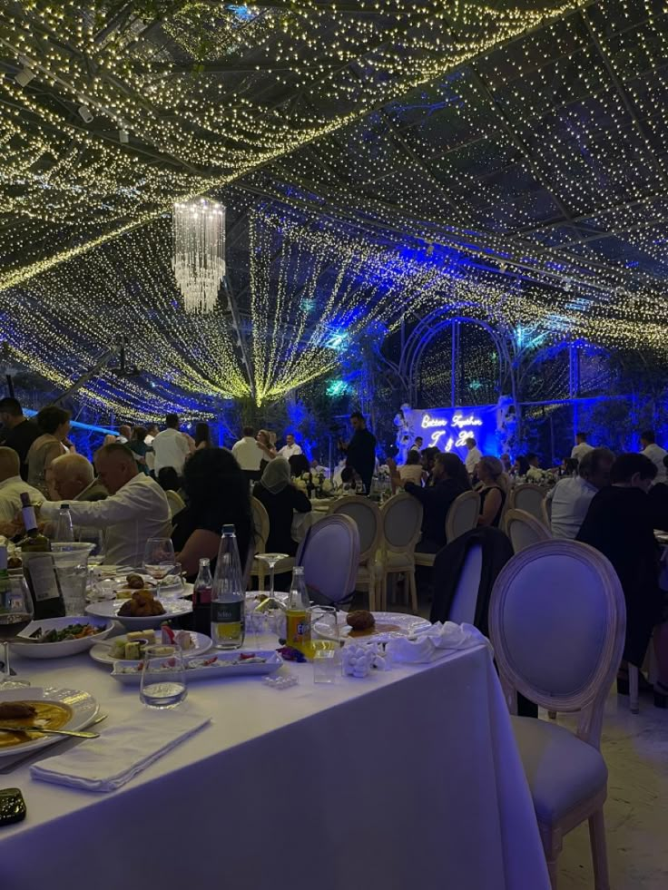 people are sitting at tables in a large room with lights on the ceiling and white linens covering the walls