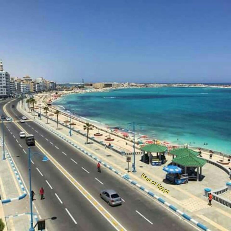 an empty street next to the ocean with cars driving on it and buildings in the background