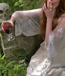 a woman in white dress holding a wine glass and talking on cell phone while sitting next to stone wall