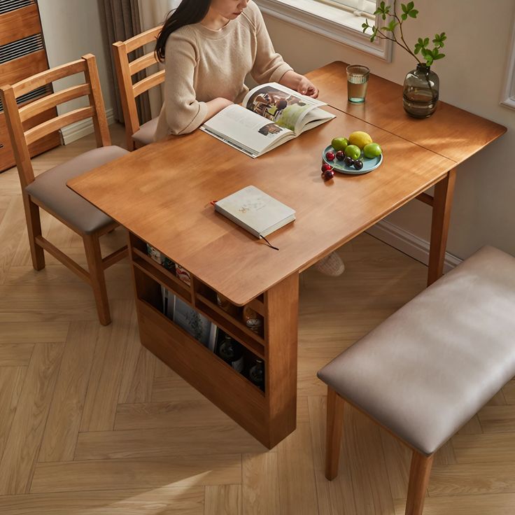 a woman sitting at a wooden table reading a book and eating fruit on the plate