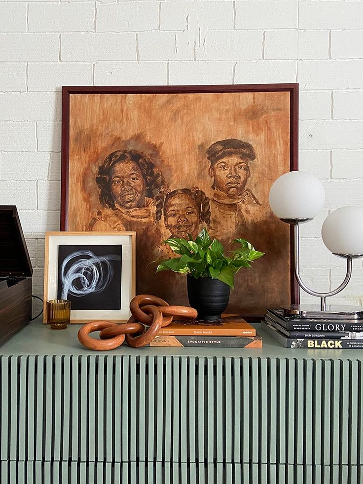 a painting and some books on a table with a plant in front of it next to a lamp