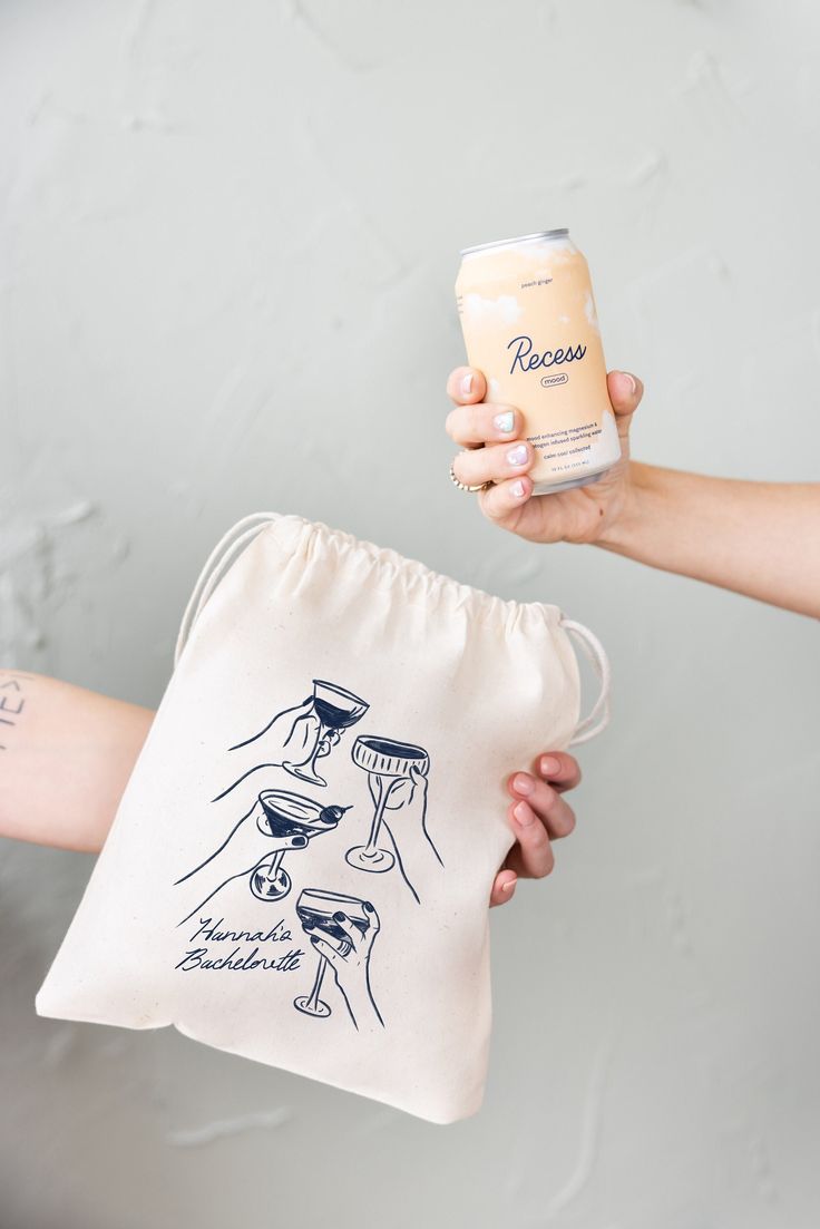 two hands holding a can of beer and a drawstring bag with drawings on it