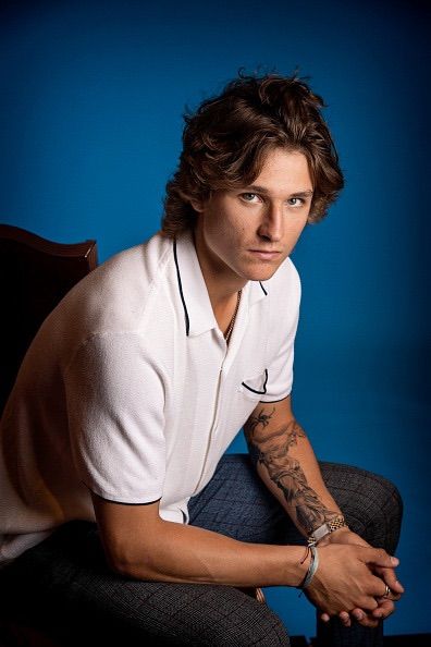 a man sitting on a chair with his arms crossed and tattoos on his arm, in front of a blue background