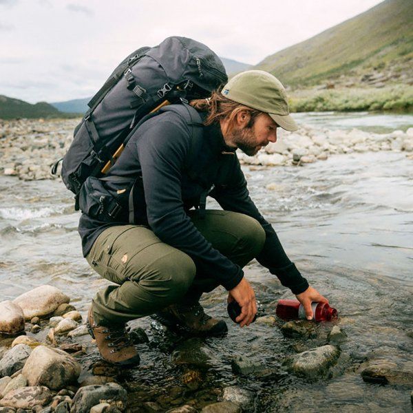 a man with a backpack and water bottle crouches down to pick up something from the river