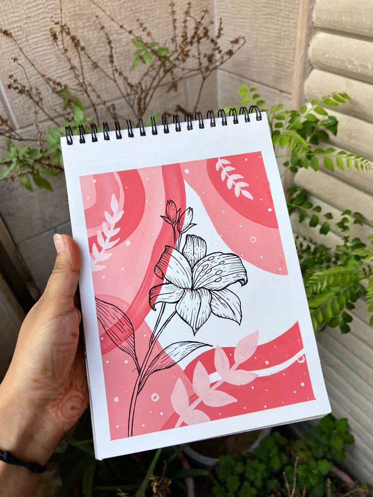 a person holding up a drawing in front of a plant with leaves and flowers on it