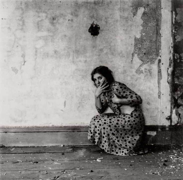 a woman is sitting on the floor in front of a wall with peeling paint and holding her hands to her face