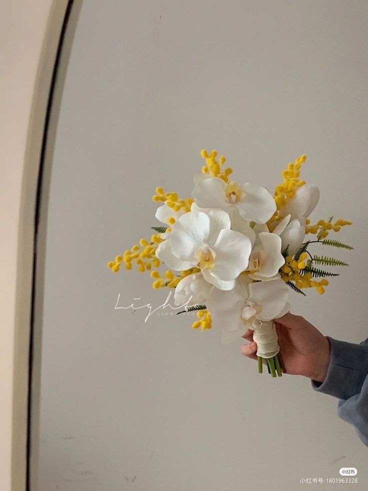 a person holding a bouquet of white and yellow flowers in front of a mirror with the reflection of their hand