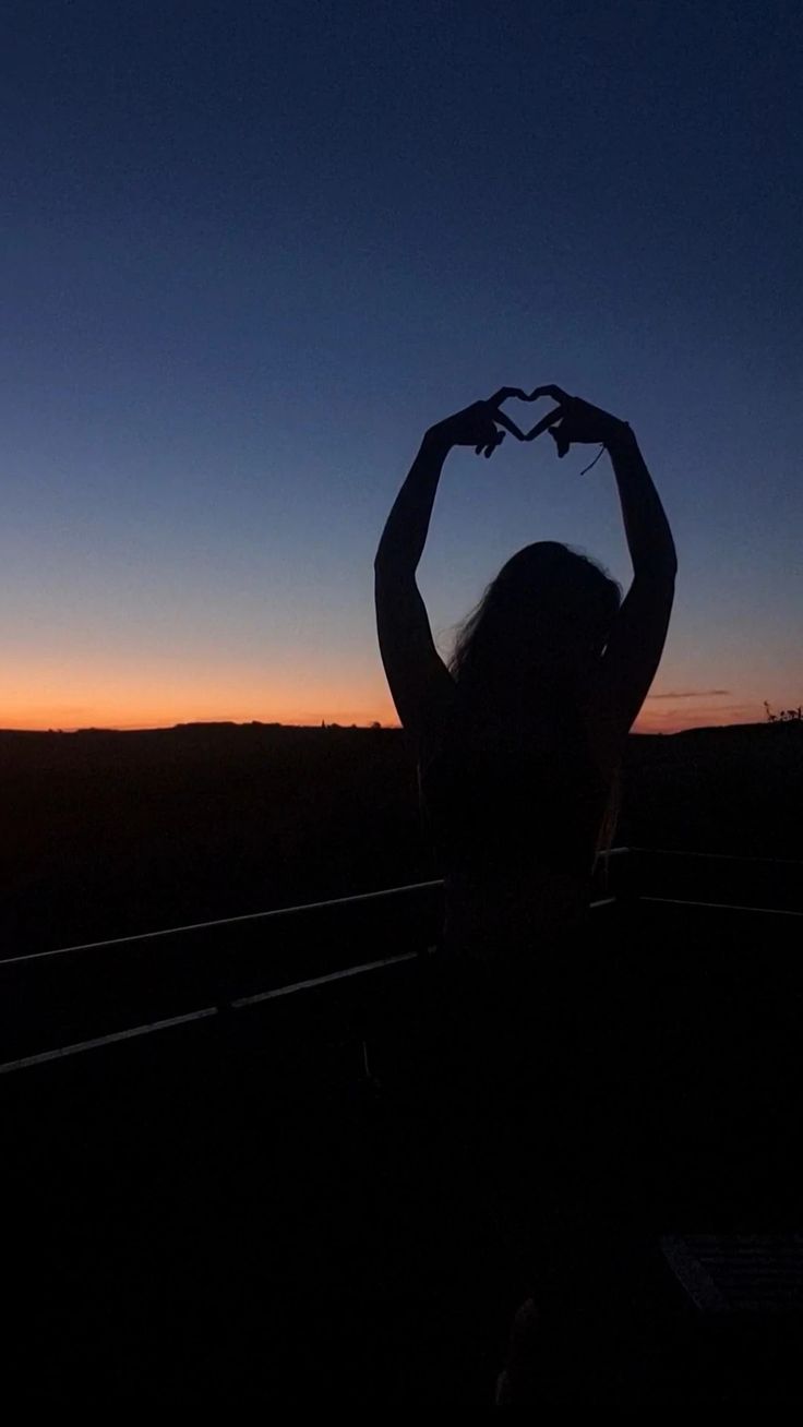 the silhouette of a woman holding her hands up in the shape of a heart at sunset