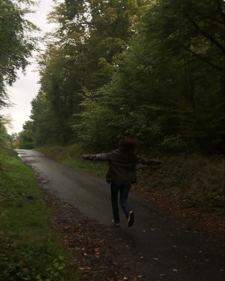 a person running down a road in the middle of some trees and grass with their arms outstretched