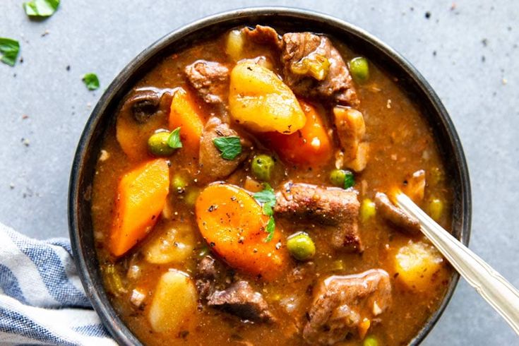 a bowl filled with stew and carrots on top of a blue towel next to a spoon