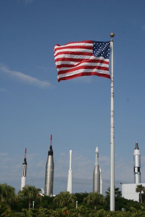 an american flag flying in the air next to some rocket like structures and trees on a sunny day