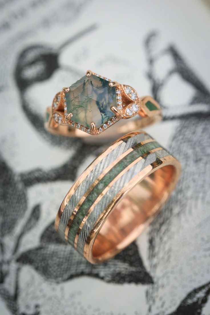 A matching wedding ring set from Staghead Designs sits on an antique bird illustration. First is our "Lucy In The Sky" design engagement ring in 14K rose gold with a hexagon cut moss agate center stone, diamond halo & accents, & moss inlays. Below is "Rio" in 14K rose gold with a centered crushed moldavite inlay & a damascus steel inlay on either side. These rings are fully customizable & can be made with a wide variety of unique materials & stones. Unique Engagement Rings Amethyst, Green Engagement Ring Set, Moss Agate Matching Rings, Unorthodox Wedding Rings, Exotic Wedding Rings, Matching Moss Agate Wedding Rings, One Of A Kind Wedding Rings, Moldavite Engagement Ring, Unique Engagement Rings Opal