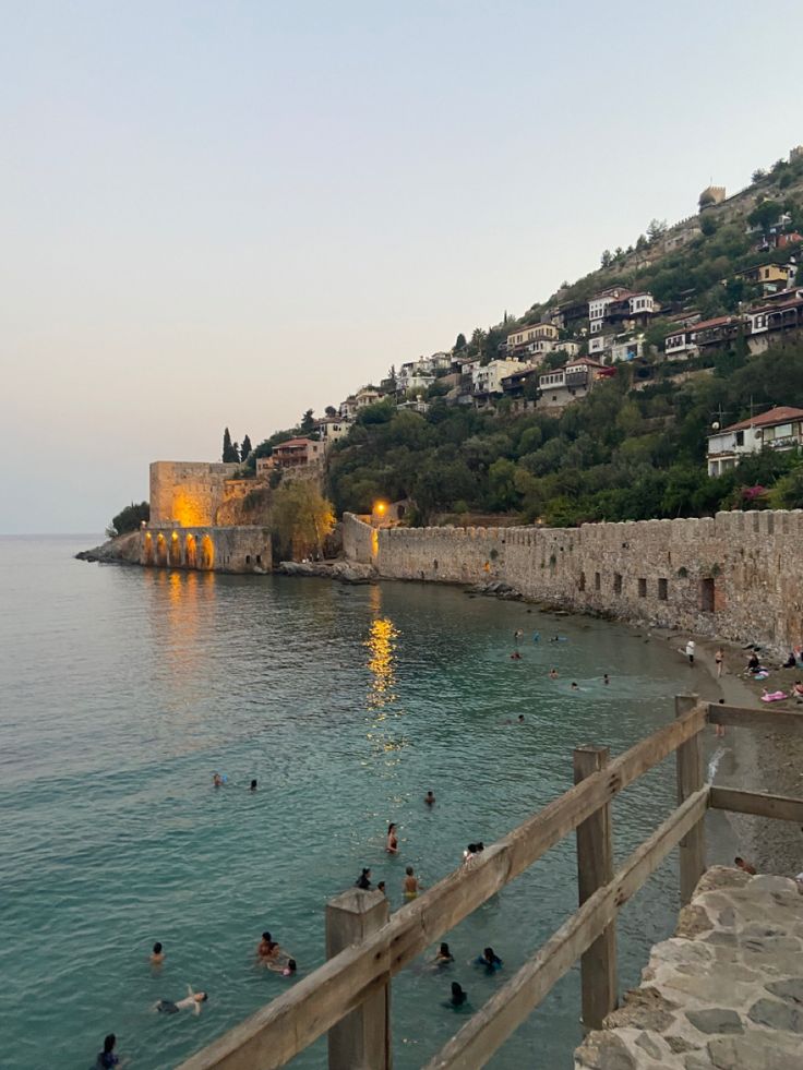 people are swimming in the water next to a stone wall and some hills with houses on it