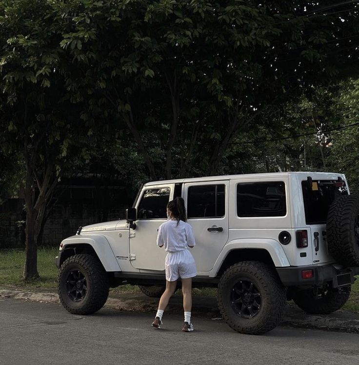 a woman walking past a white jeep parked on the side of a road next to trees