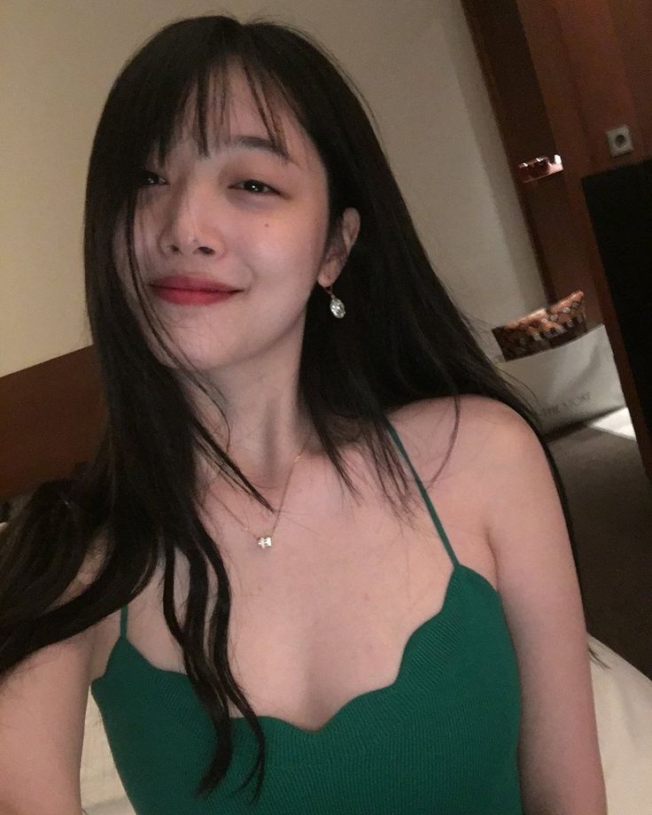 a woman with long black hair wearing a green top sitting on a bed in front of a mirror