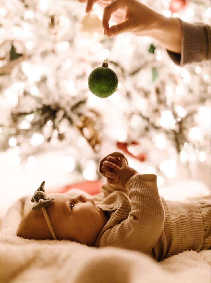 a baby laying on its back in front of a christmas tree with a hand reaching for an ornament