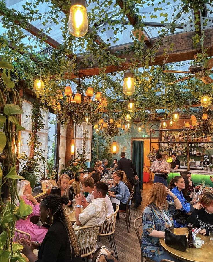 many people are sitting at tables in a restaurant with plants growing on the ceiling and hanging lights above them