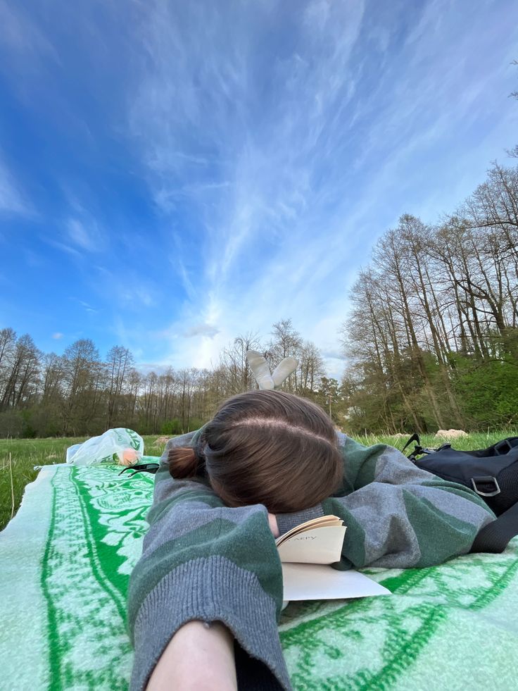 a woman laying on top of a green blanket next to a lush green forest under a blue sky