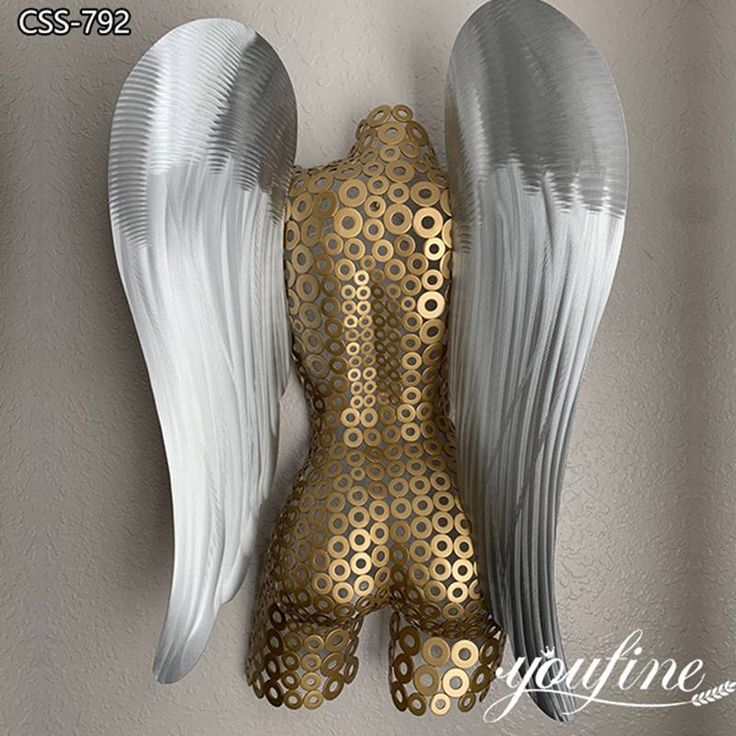 an angel statue with gold and white wings hanging on the wall in front of a gray background