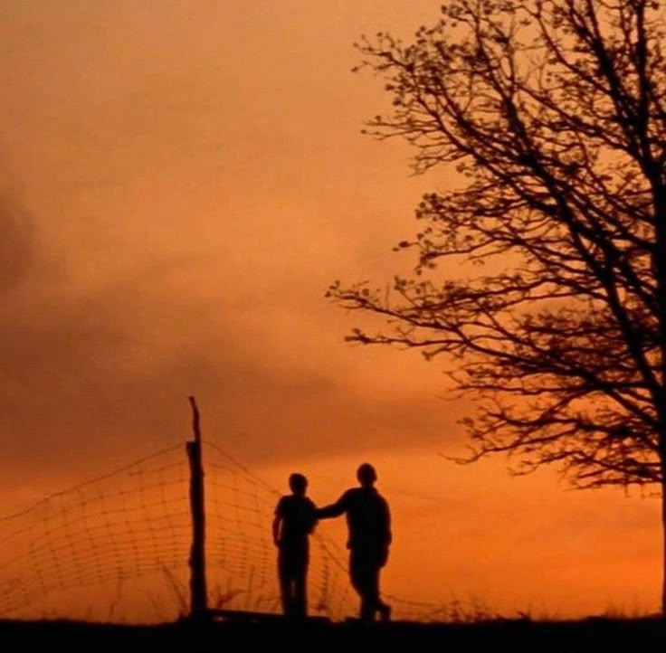 two people standing next to each other in front of a fence and tree at sunset