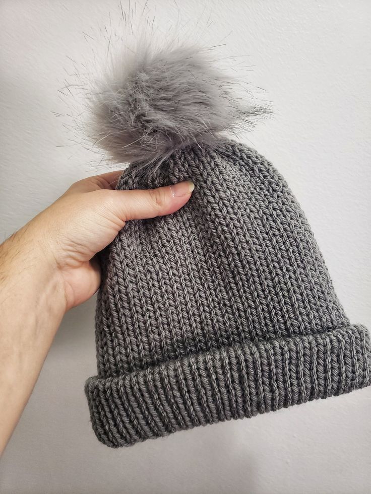 a hand holding a gray knitted hat with a grey pom - pom