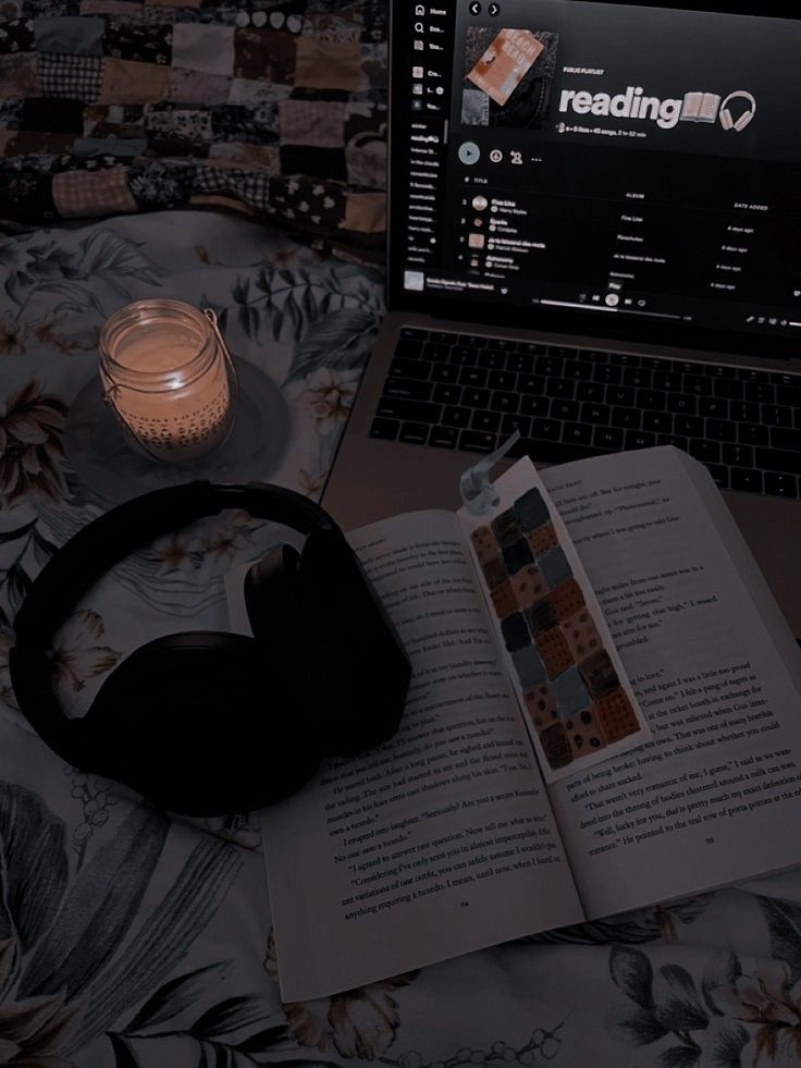 an open book, headphones and candle sit on a bed next to a laptop