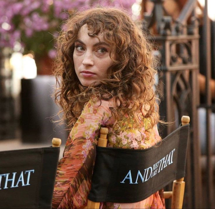 a woman with curly hair sitting in a chair and looking off to the side while holding her arm over her shoulder