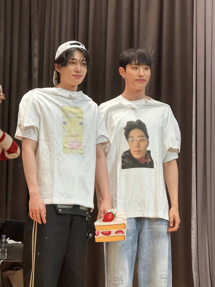 two young men standing next to each other in front of a curtain holding sandwiches and fruit