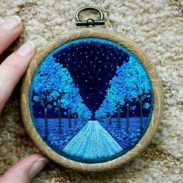 a hand holding a small embroidered ornament with trees and road in the background