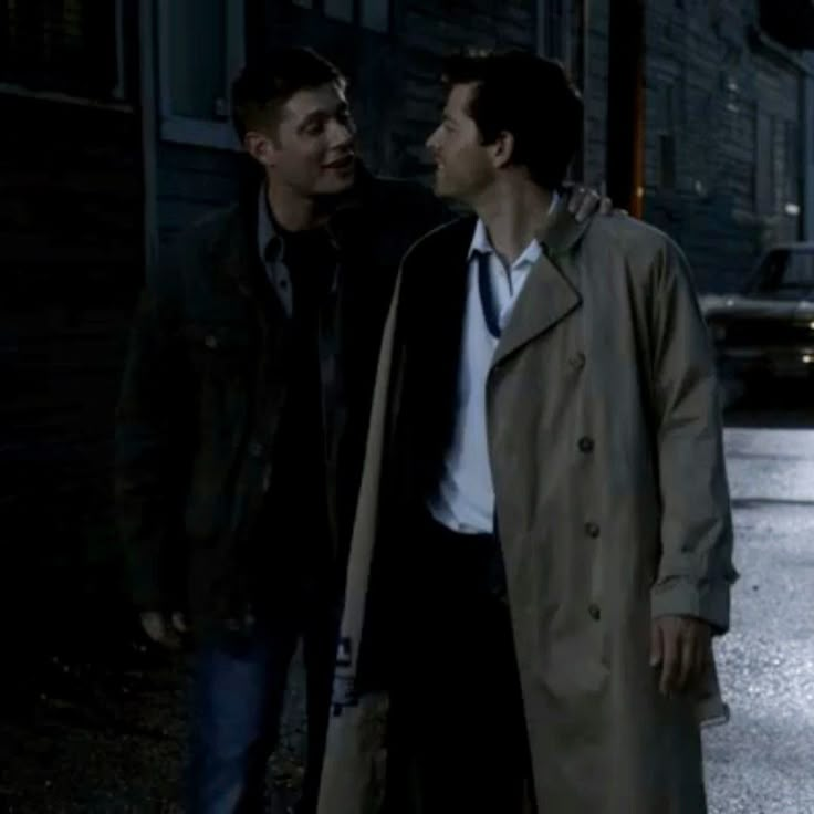 two men standing next to each other in the street at night, one wearing a trench coat