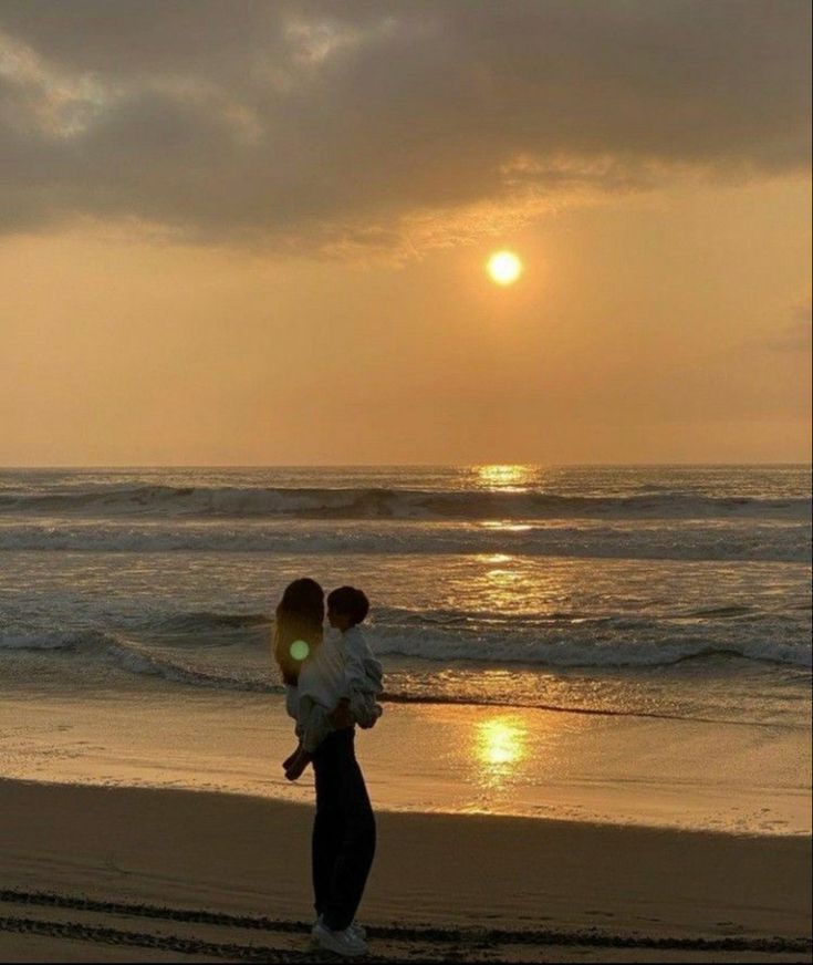 two people walking on the beach at sunset, one carrying a child in his arms