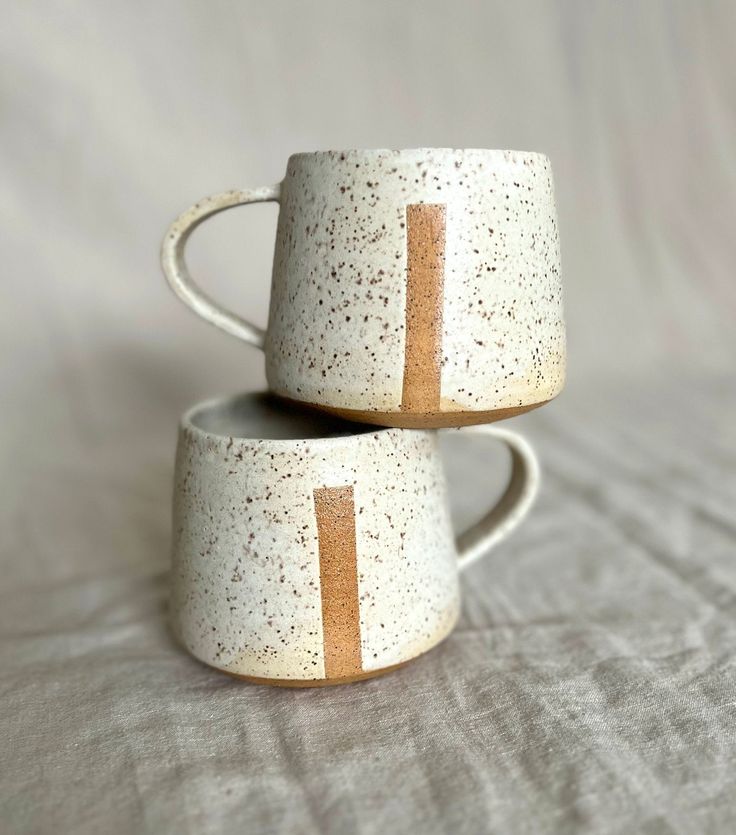 two white cups with gold lines on them sitting on a bed sheet, one has a brown stripe in the middle