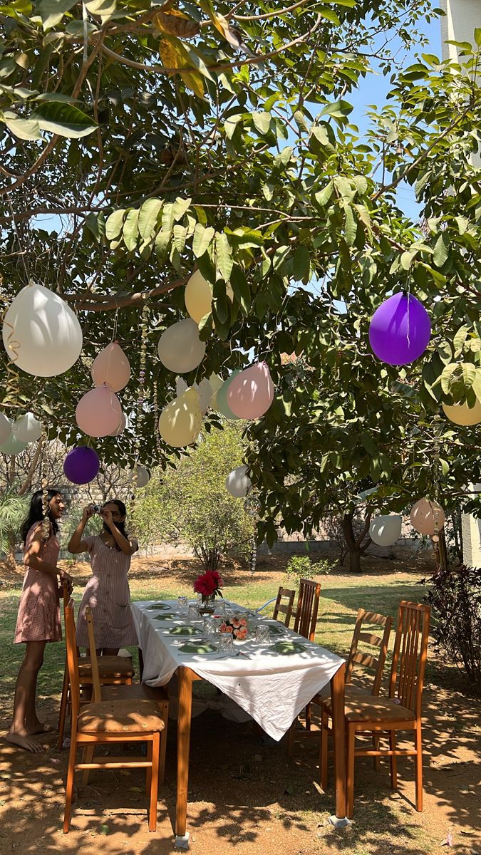 two women sitting at a table under some paper lanterns hanging from a tree with purple and white balloons