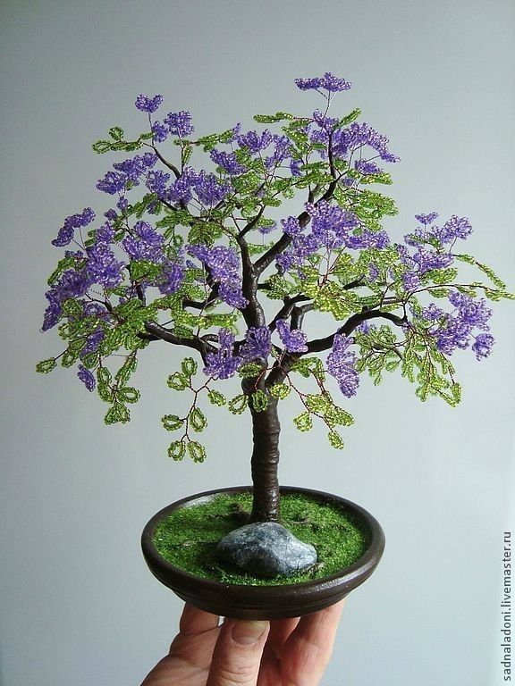 a hand holding a miniature tree with purple flowers and green leaves on it's base