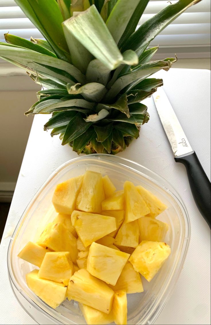 sliced pineapple in a plastic bowl next to a knife on a white tablecloth