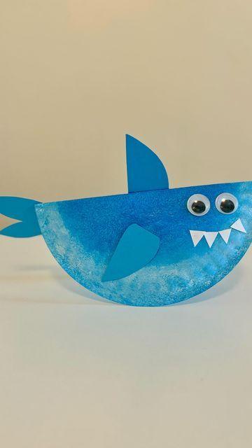 a paper plate shaped like a blue shark with big eyes and sharp teeth, sitting on a white surface