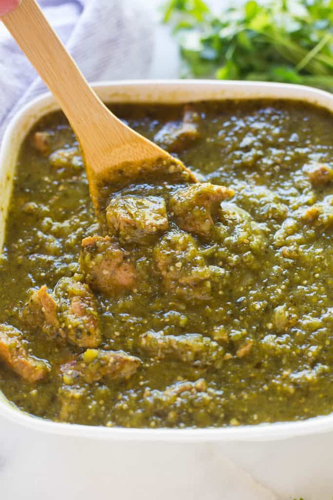 a wooden spoon in a white bowl filled with green sauce and meatballs on the side