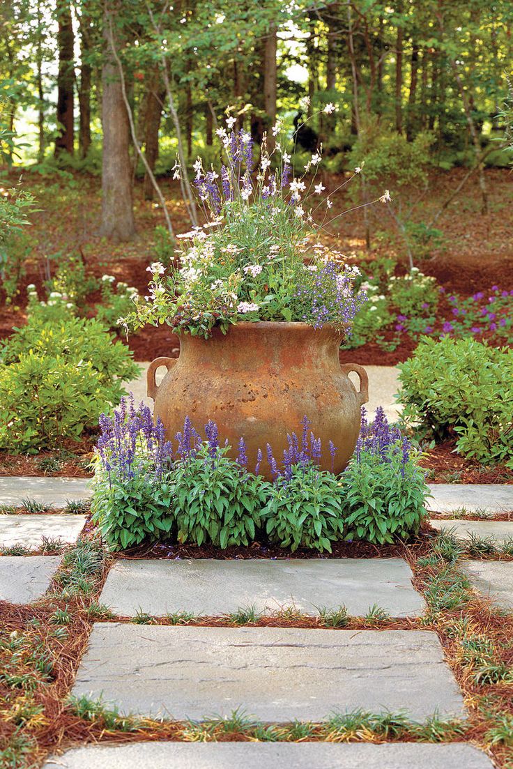 a large vase sitting on top of a stone walkway surrounded by flowers and greenery