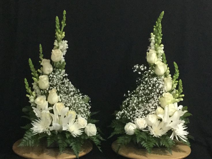 two vases with white flowers and greenery on wooden bases against a black background