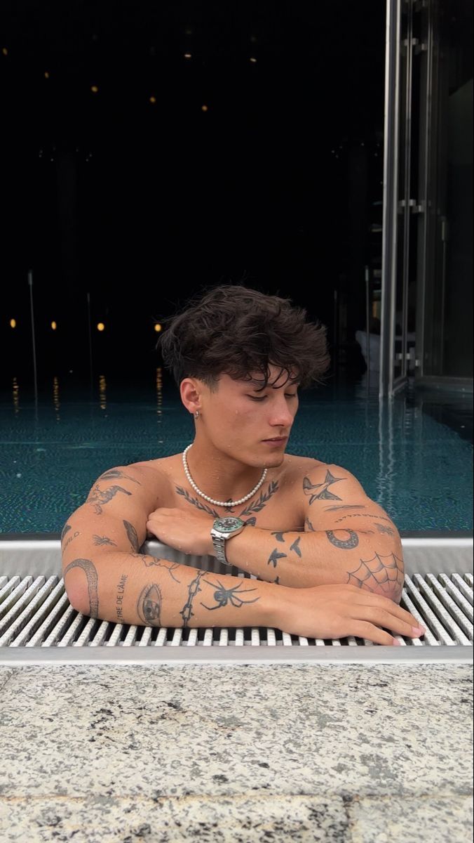 a man with tattoos on his body sitting next to a swimming pool and looking at the camera