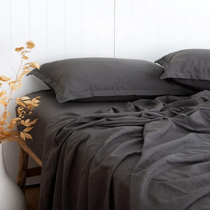 a bed with grey sheets and pillows next to a white vase filled with flowers on top of it