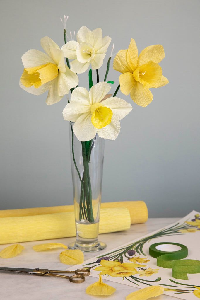 yellow and white flowers in a clear vase on a table with paper, scissors and tape