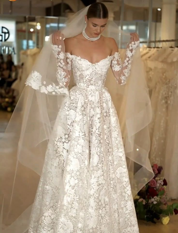 a woman wearing a wedding dress with long sleeves and a veil on her head standing in front of some dresses