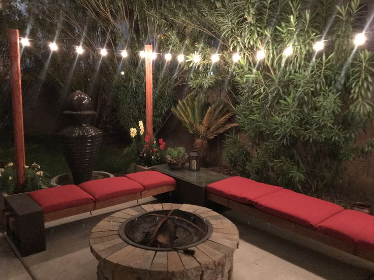 an outdoor fire pit with red cushions and lights on the trees in the back yard